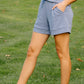 Woman in a sleeveless top and shorts standing on a grassy field with trees in the background